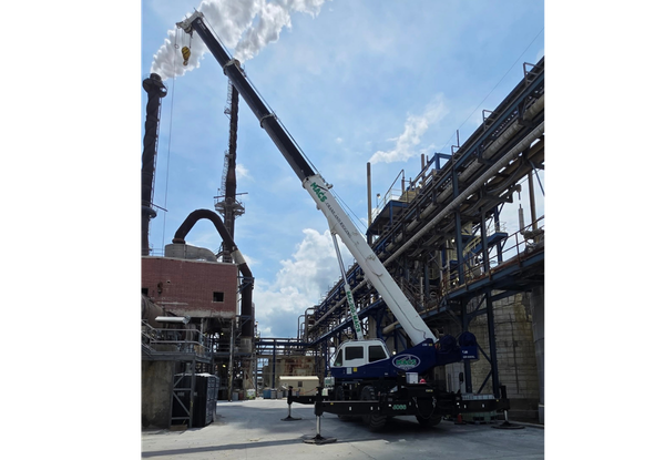 Mobile crane lifting a vertical pipe at an industrial construction site, with metal scaffolding and tall equipment around.